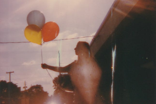 A worn polaroid of a woman holding up balloons to represent distant childhood memories and observing something lost.