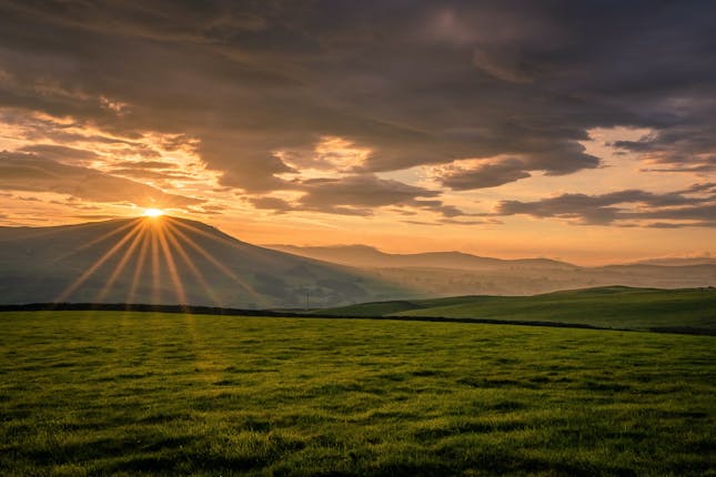 sunlight piercing through clouds above a green field and mountain representing the beauty of creation 