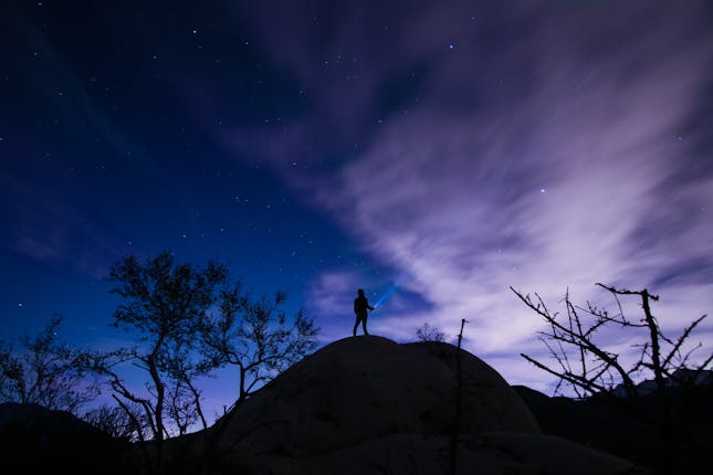 A man atop a hill shining his flashlight into the night sky - representing the search for truth about God.